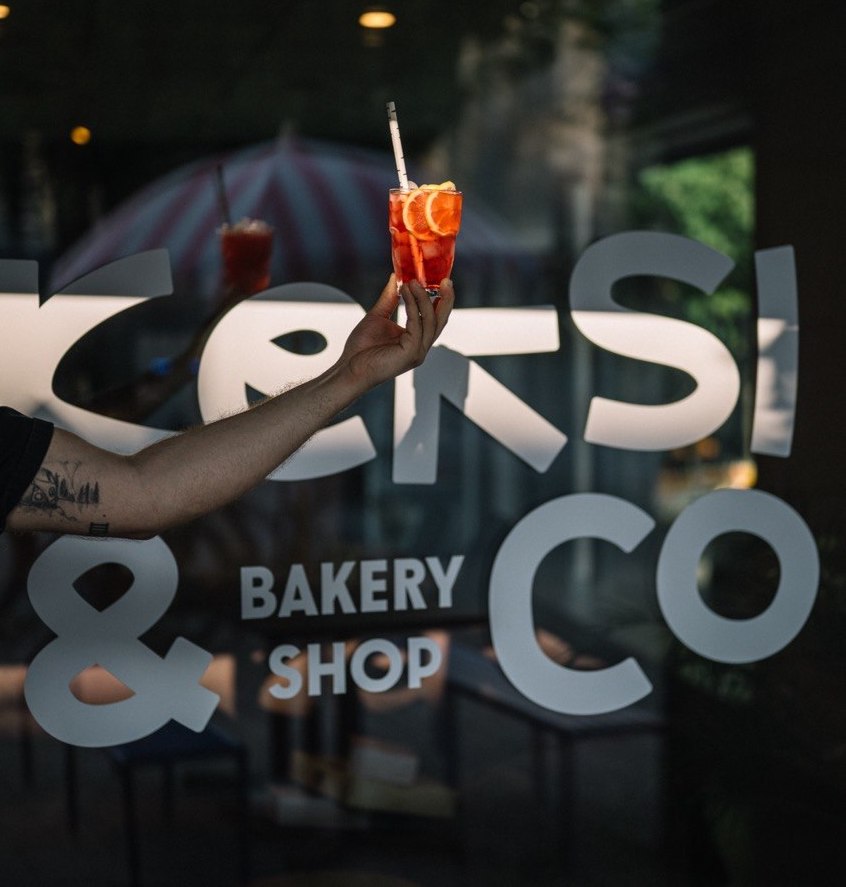 A hand raising a drink in front of the Keksi & Co bakery shop window